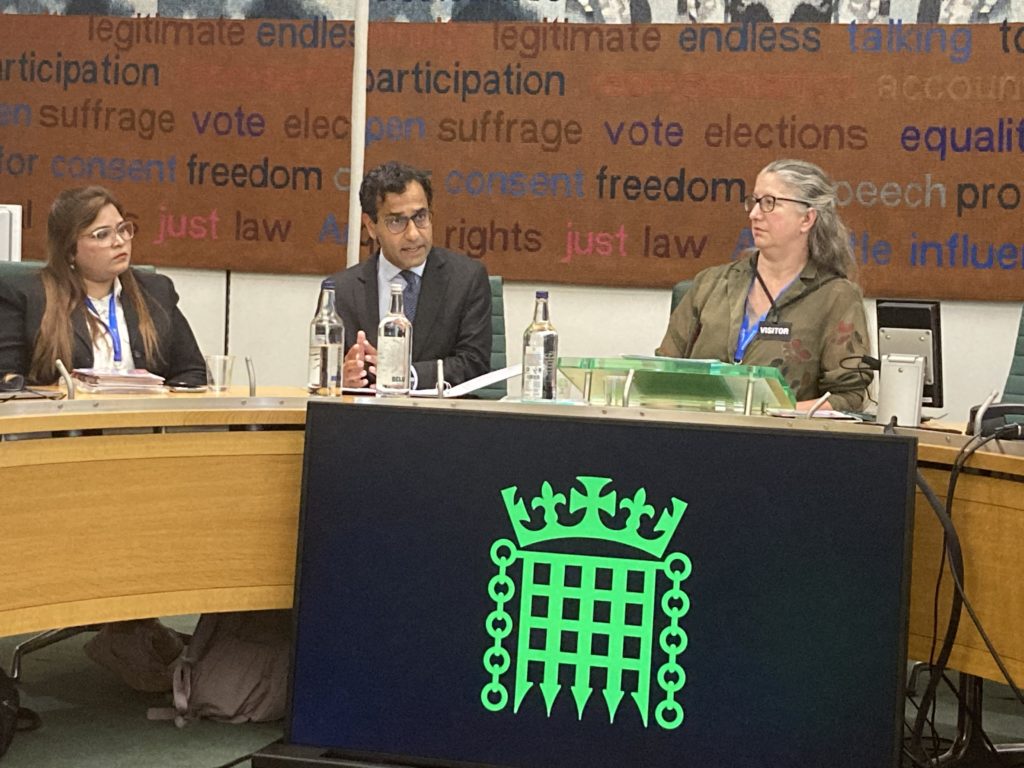 Photograph of a man speaking in UK parliamentary committee room, with two women sat next to him