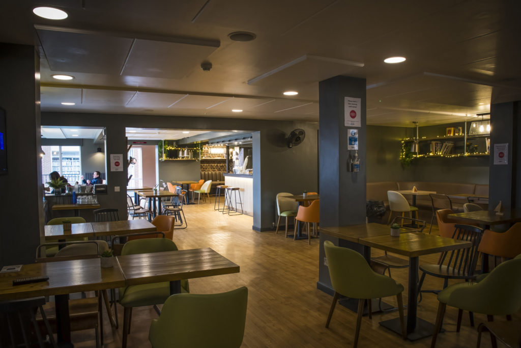 The main area of the IDS Bar and Kitchen. The angle the shot is taken from does not show the main canteen, but shows the majority of the seating and tables, with space in the middle. In the background, the coffee and drinks bar can be seen, as well as some visitors in the second part of the bar and kitchen. There is a square archway between the two parts of the bar.