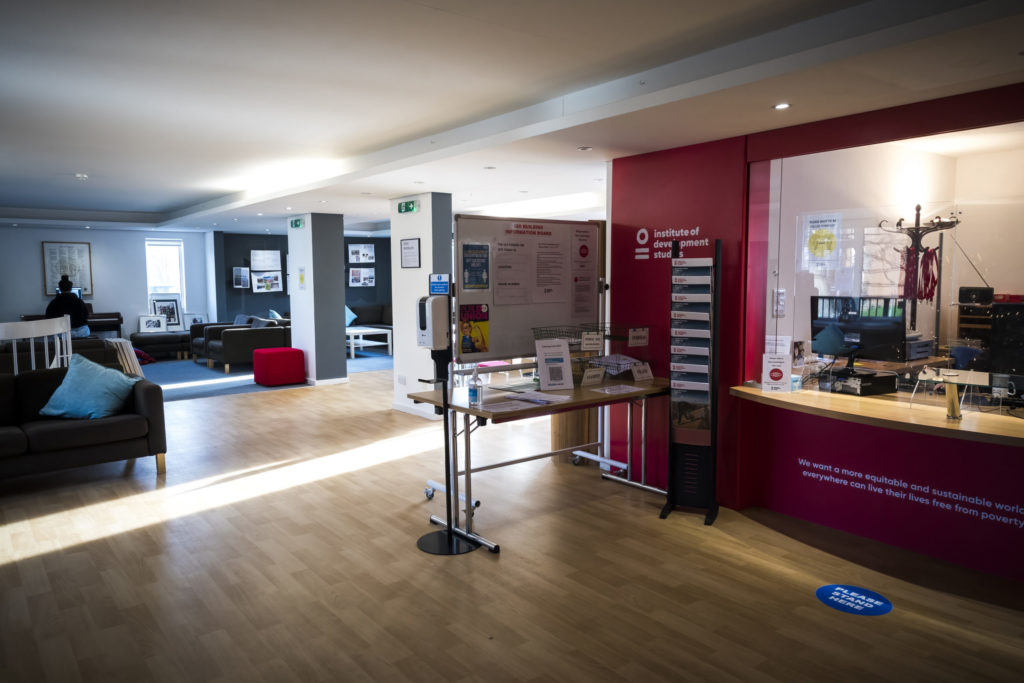 The IDS main reception area. The shot is taken diagonally so that the reception desk is to the right hand side of the photo. The floor is smooth wood effect. The wall around the reception glass is painted in the IDS brand red. In the background the couch can be seen and behind that, the seating and pillars to the left hand side of the IDS reception.