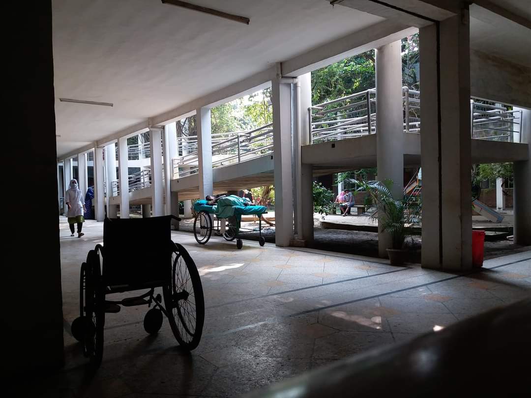 In the foreground there is an empty wheelchair facing away from the camera. It sits in the outdoor corridor of a hospital that looks onto a courtyard with trees. In the corridor there is also a person lying on a hospital bed and a medical professional walking down the corridor.
