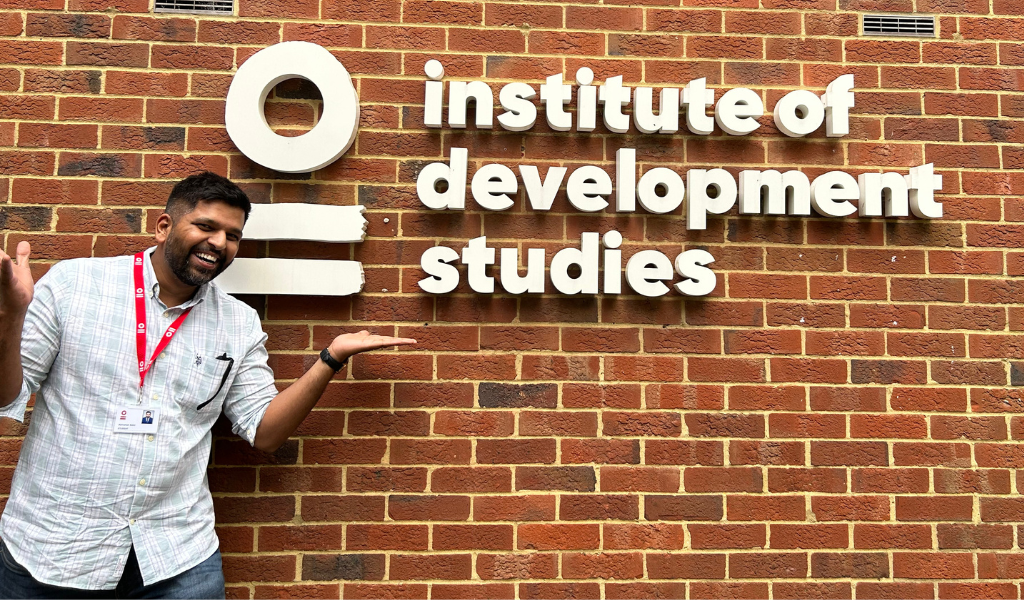 An Indian man in a white shirt wearing a red lanyard stands in front of a brick wall, with the words 'Institute of Development Studies' written in front of it.