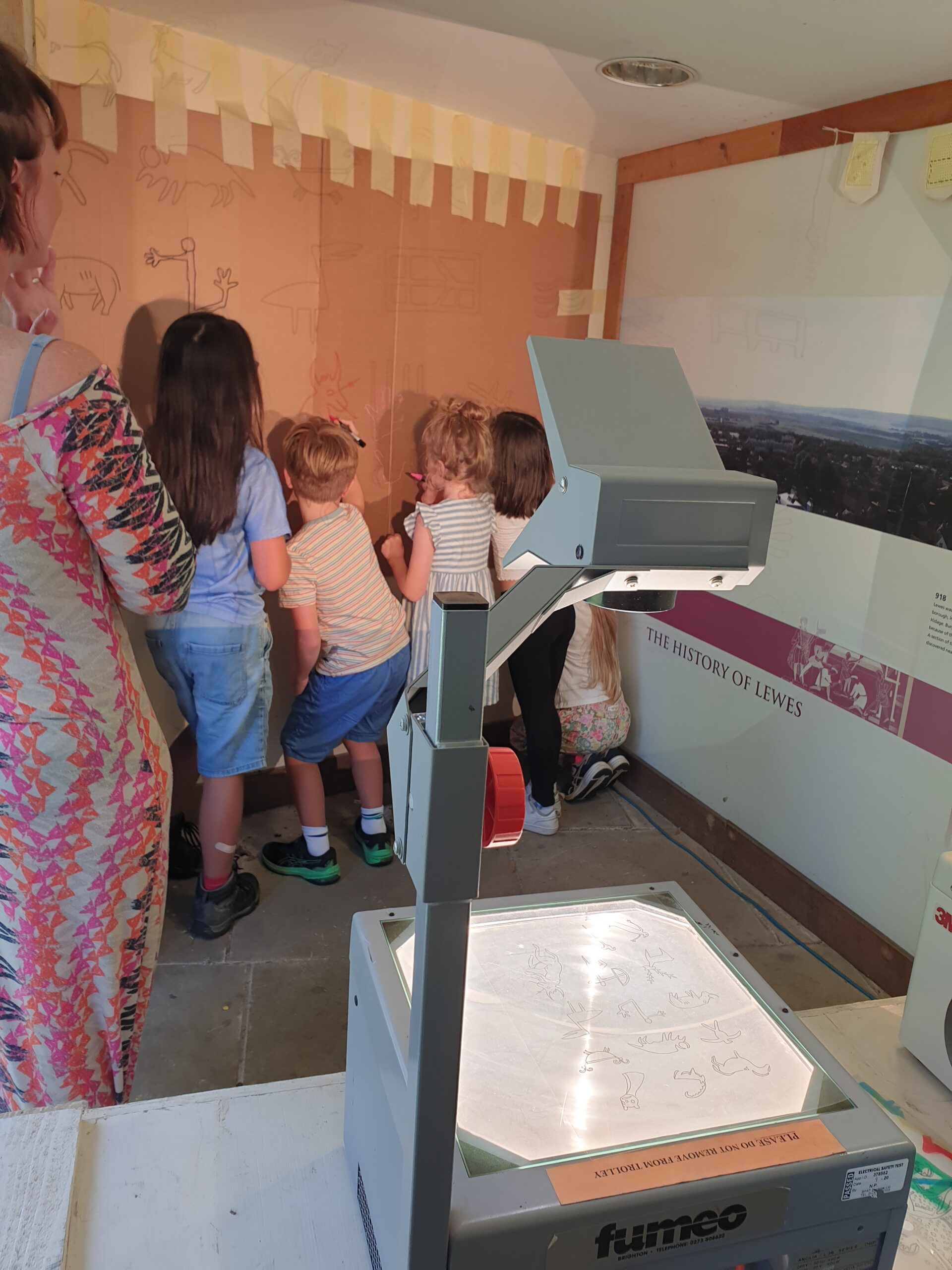 Four children draw on a wall where a big peice of paper has been stuck. There are shapes being projected onto the wall for the children to add to, from an overhead projector. An adult watches the children. On another wall it reads "The history of Lewes"