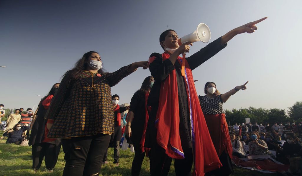 A film still from This Stained Dawn depicting a group of women wearing black and red in formation pointing offscreen. Behind the, a mostly female audience sits on the grass and watches the performance 