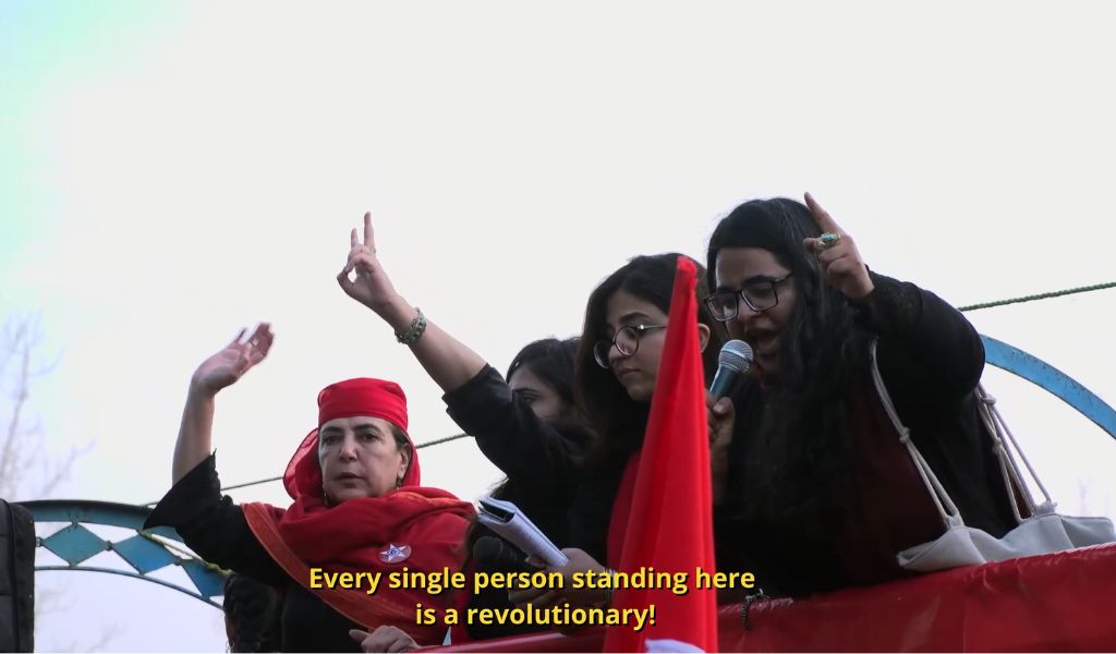 A film still from This Stained Dawn depicting four women inblack and red looking down at a crowd from a pick up truck. One of them holds a mic, and is speaking with her finger pointing at the sky. Another woman holds up a sign. An older woman has her head bandaged with a red scarf