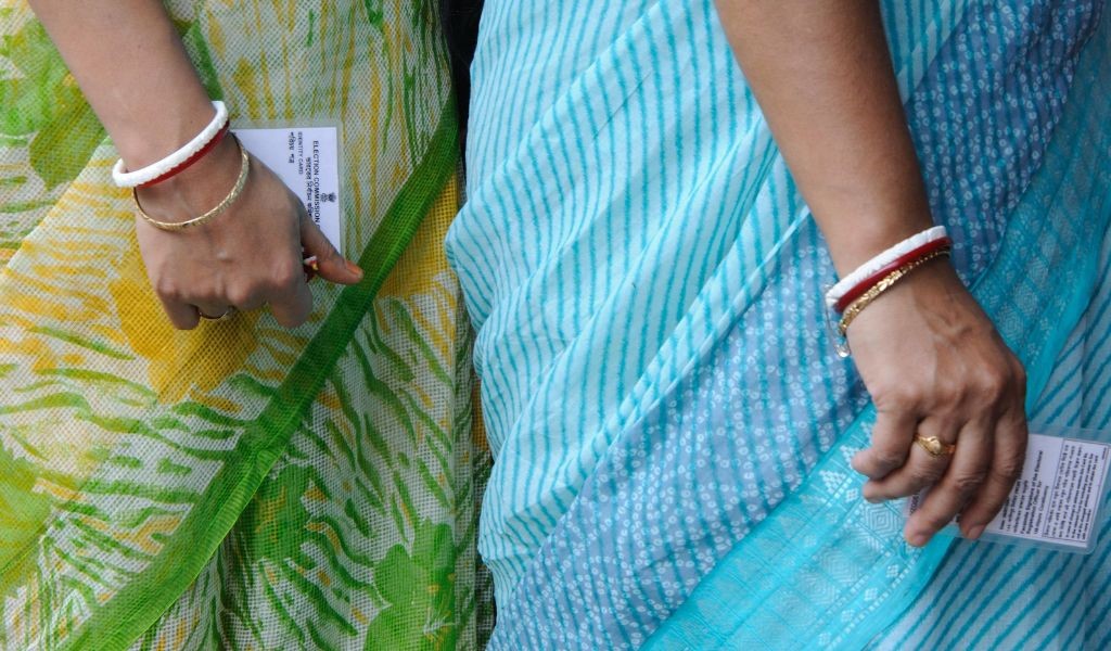 Close up of two women's hands down by their sides holding election voting cards. They are wearing bangles on their wrists and blue and green saris.