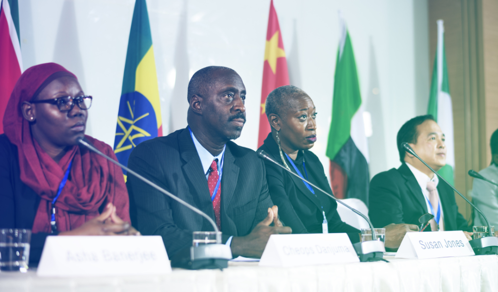 Four people sit in a line at a high level meeting, with microphones in front of them and world flags in the background.