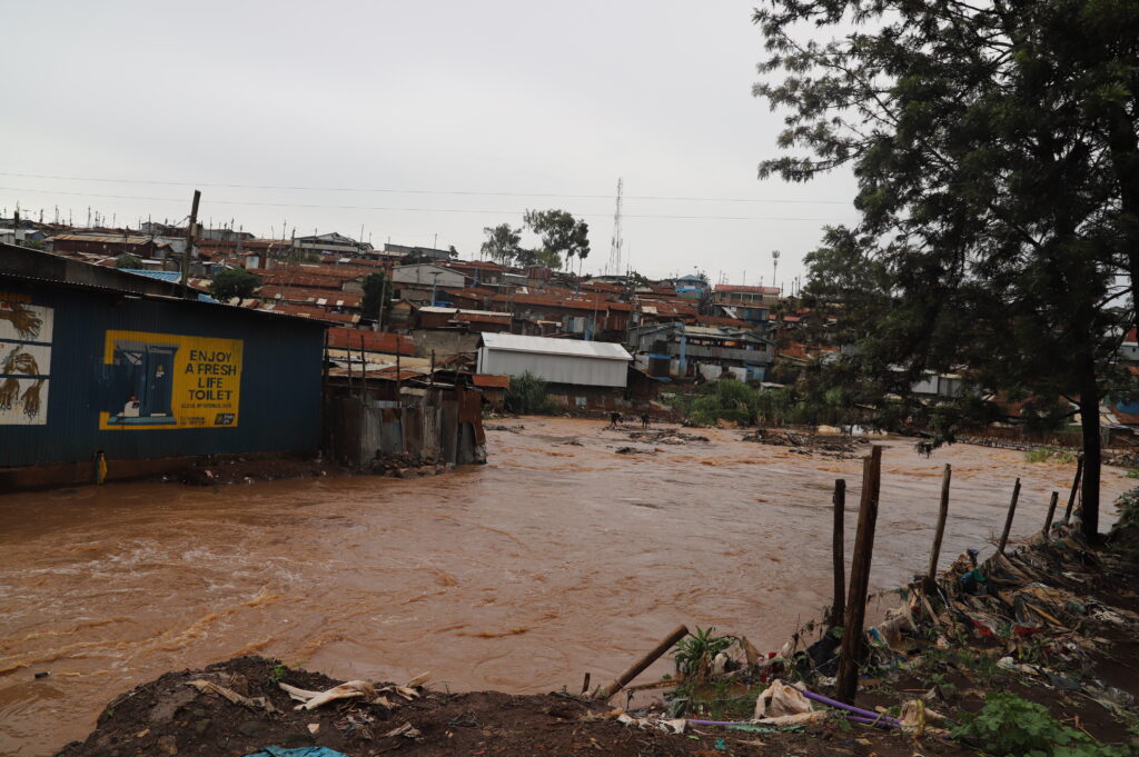 A photo of a flooded river flowing through an urban area in Nairobi, Kenya, on a rainy day. There are many buildings on the far side of the river.
