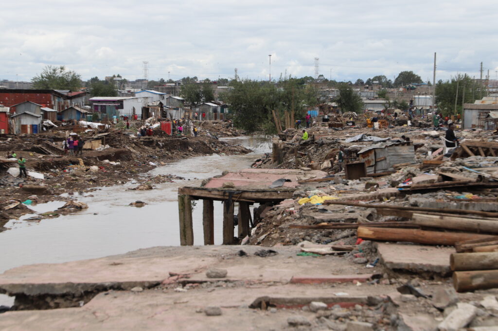 A photo showing he aftermath of flooding in an urban area or Nairobi, Kenya. There is a small river running from the bottom left to the right of the photo. Destroyed buildings are everywhere, with lots of mud and metal.