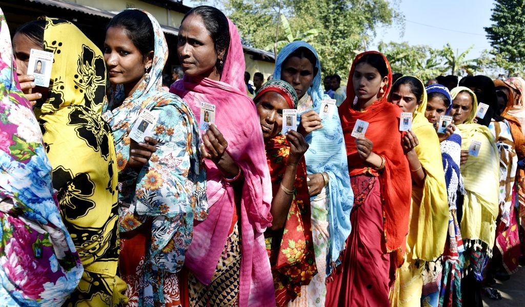 Women in colourful saris queue up in a line in the sun holding up ID cards for voting purposes. The tops of trees can be seen in the background.