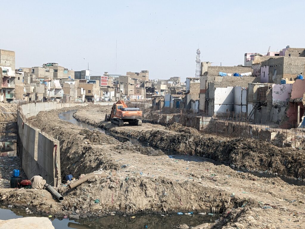 A photo of a raised waterway running through an urban area of Karachi, Pakistan. There are concrete buildings on either side of the water way, which itself is surrounded by dry earth. There is an orange construction vehicle next to the water way.
