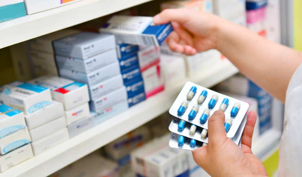 A close up of a person holding packets of pill capsules with their left hand and with their right hand putting a box of medication on a shelf full with piles of other boxes of different medication.