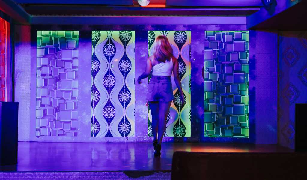 A young woman dressed in short denim shorts and a white top walking to the back of a stage in a night club. The stage is lit blue.