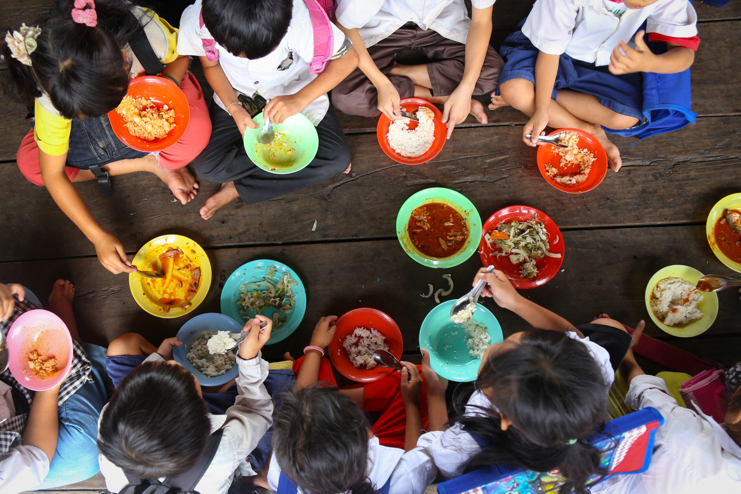 Overhead view of eight children sitting on a dark wooden floor in a circle, each eating from a colourful bowl in front of them. They all have dark hair, wearing white shirts and rucksacks on their back.