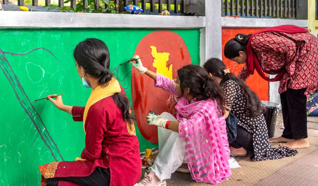 Four women painting murals in bright green, red and yellow colours on a wall outdoors. They are sitting on the ground facing the wall with paint pots and brushes in hand.